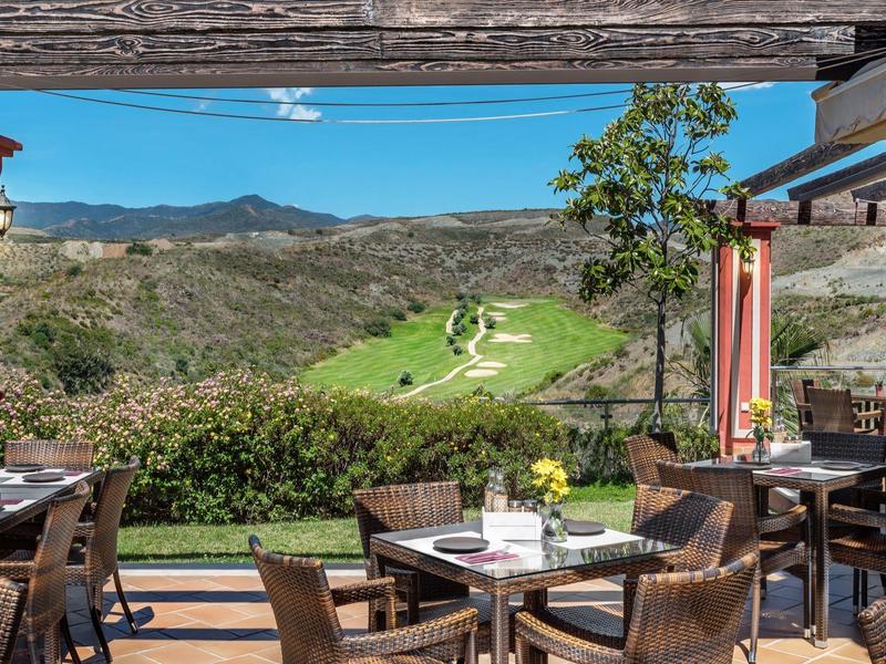 Terrasse mit Stühlen und Tischen, Blick auf grüne Hügel und blauen Himmel im Hintergrund.