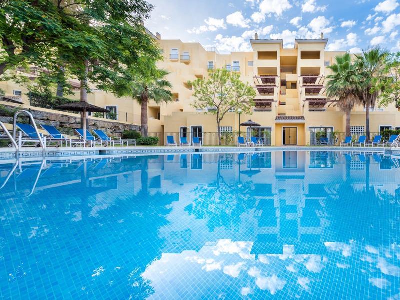 Großer blauer Pool neben mehrstöckigem Hotel mit Palmen und blauem Himmel mit Wolken.