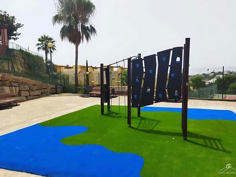 Outdoor-Fitnesspark mit blauer und grüner Gummimatte, schwarzen Trainingsgeräten und Palmen im Hintergrund.