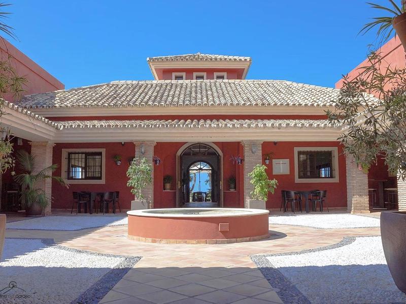 Innenhof eines Hauses mit roter Fassade, Springbrunnen in der Mitte, blauem Himmel und Pflanzen.
