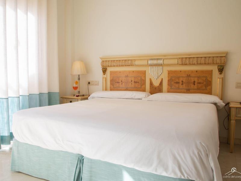 Schlichtes Schlafzimmer mit großem Doppelbett, hellem Holzrahmen und weißen Vorhängen.