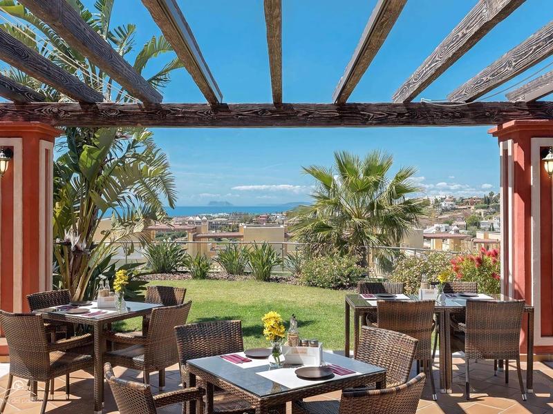 Terrasse mit Tischen, Stühlen, Palmen und Blick auf Stadt und Meer unter blauem Himmel.