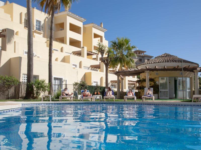 Schwimmbad vor einem hellen Hotelgebäude mit Palmen und Sonnenschirmen bei blauem Himmel.