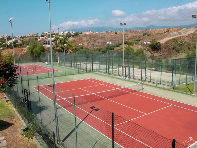 Leerer Tennisplatz mit rotem Spielfeld und grüner Umrandung, umgeben von Maschendrahtzaun.