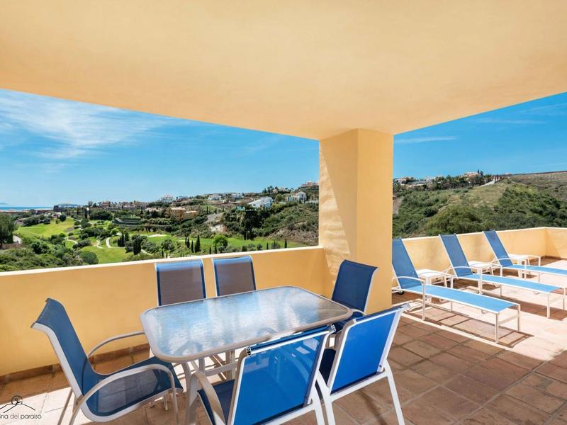 Große Terrasse mit blau-weißen Stühlen, Glas-Tisch und Liegen, Blick auf grüne Hügel.