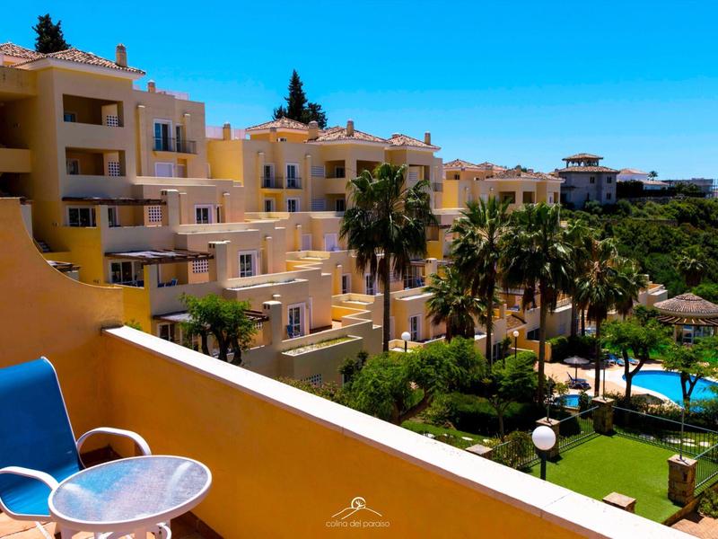 Mehrstöckiges gelbes Gebäude mit Balkon, Palmengarten und blauem Pool unter klarem Himmel.