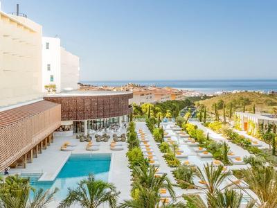 Hotelgarten mit Swimmingpool, Palmen und Sitzgelegenheiten, dahinter das blaue Meer und Strand.