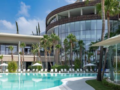 Moderner Hotelpool mit Sonnenliegen und Palmen bei sonnigem Himmel.