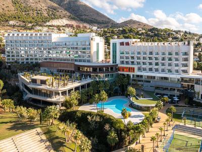 Moderner Hotelkomplex mit Pool, Tennisplatz und grünen Gartenflächen vor Bergkulisse unter blauem Himmel.