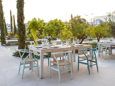 Área moderna de restaurante al aire libre con mesas blancas y sillas coloridas entre árboles.