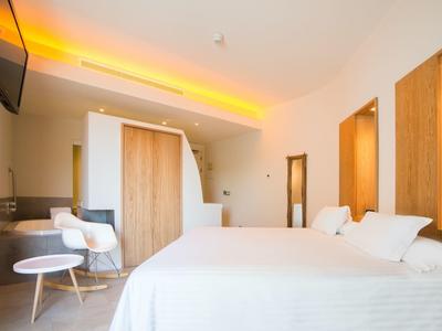 Helles Schlafzimmer mit großem weißen Bett, Holzvertäfelung, zwei Sesseln und warmem Deckenlicht.