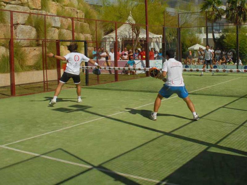 Zwei Männer spielen Paddle auf einem grünen Platz mit Zaun und Zuschauern im Hintergrund.