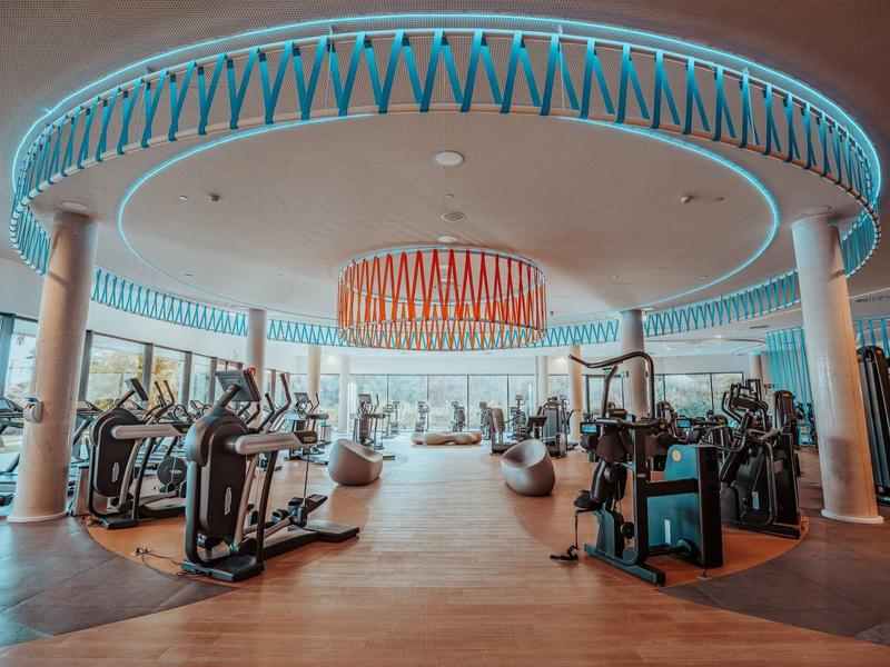 Moderner Fitnessraum mit Geräten, Holzfußboden, großen Säulen und dekorativer Deckenbeleuchtung.