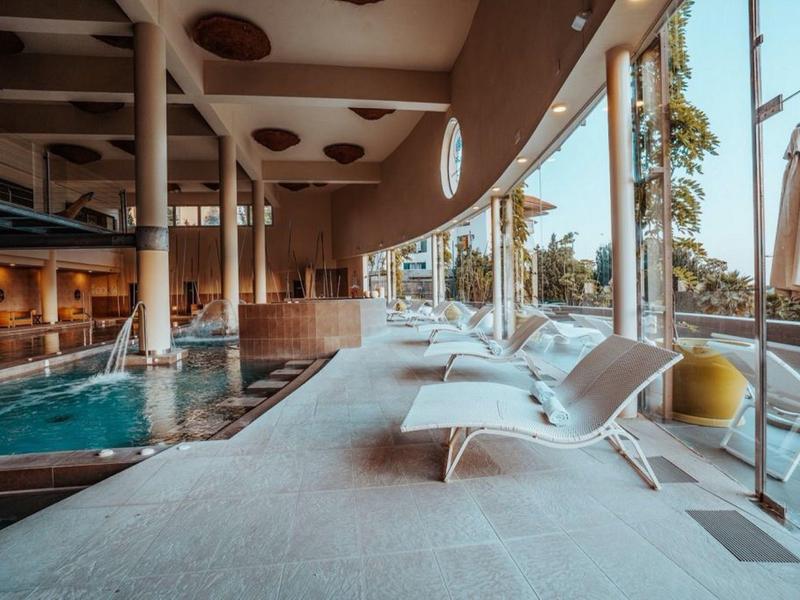 Moderner Innenpool mit großen Glasfenstern, weißen Liegen und Säulen entlang des Beckens.