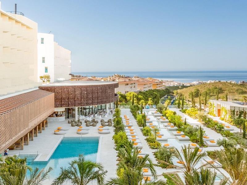 Hotelgarten mit Swimmingpool, Palmen und Sitzgelegenheiten, dahinter das blaue Meer und Strand.