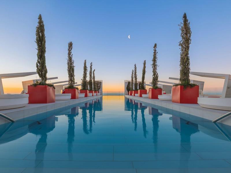 Symmetrischer Pool mit hohen Bäumen, roten Pflanzgefäßen und klarem Himmel im Sonnenuntergang.