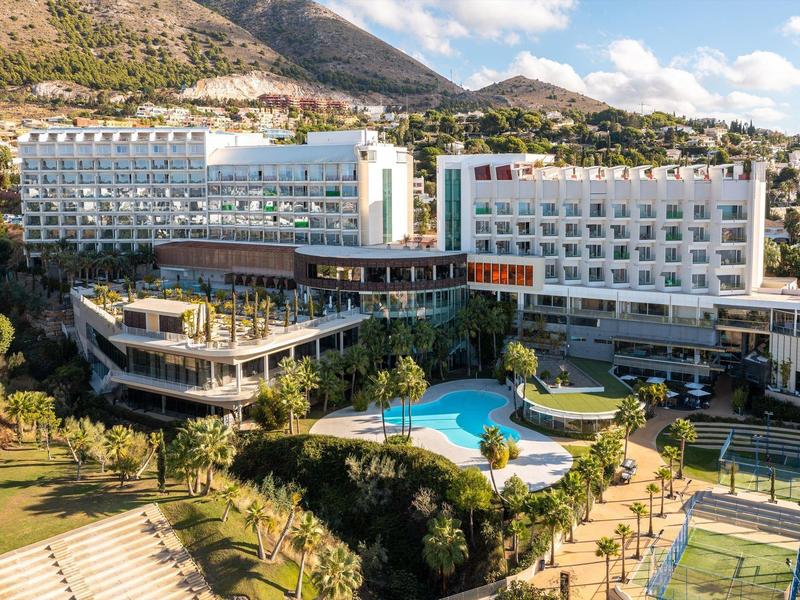 Moderner Hotelkomplex mit Pool, Tennisplatz und grünen Gartenflächen vor Bergkulisse unter blauem Himmel.