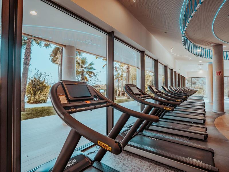 Fitnessraum mit Laufbändern entlang großer Fenster mit Blick auf Palmen und Pool