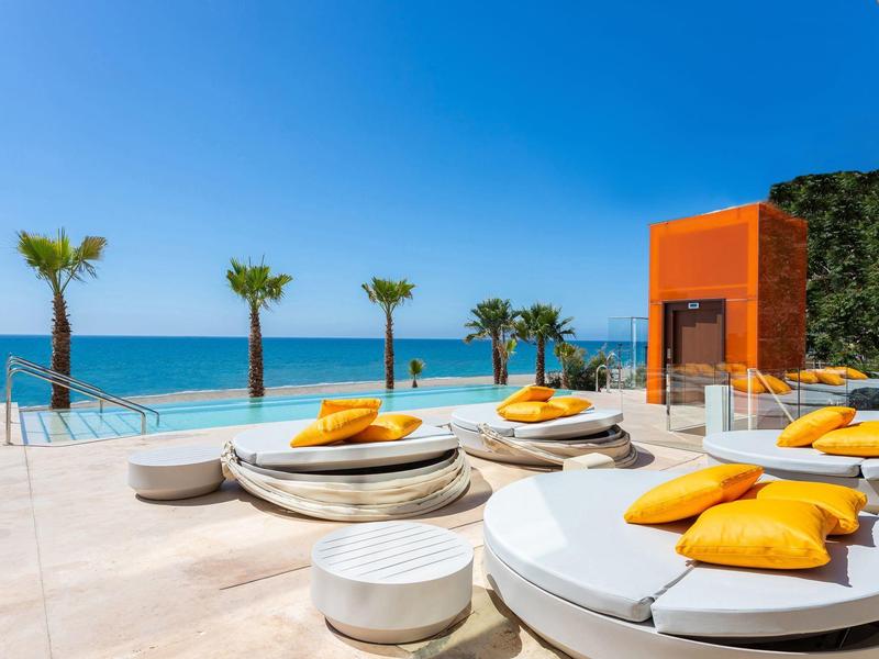 Luxuriöse Sonnenterrasse am Meer mit runden Liegen, gelben Kissen und Palmen unter blauem Himmel.