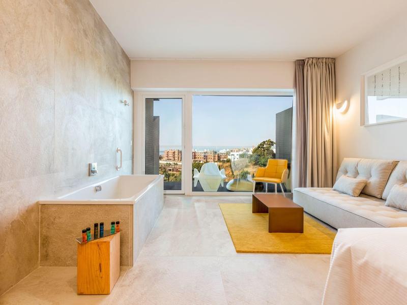 Modernes Hotelzimmer mit Badewanne, Sofa, gelbem Teppich und Blick auf die Stadt durch große Fenster