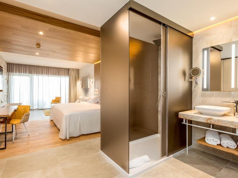 Modernes Hotelzimmer mit Dusche in der Mitte, braunen und weißen Farbtönen und großen Fenstern.