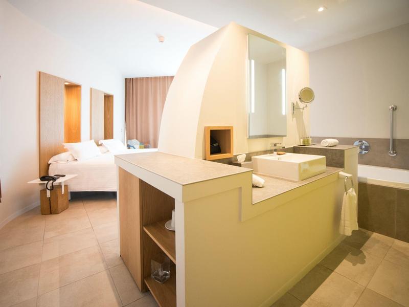 Modernes Hotelzimmer mit Doppelbett, Kamin, Badewanne und Waschtisch in hellem Design.