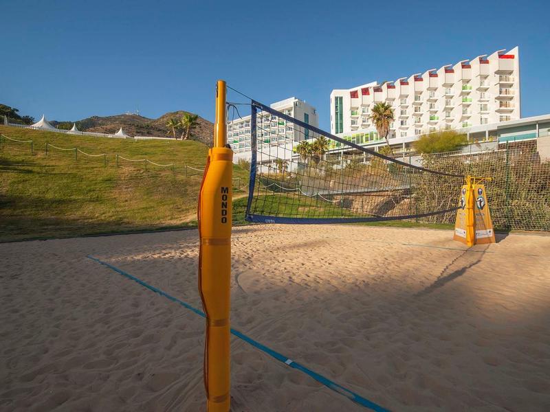Sandvolleyballfeld mit gelben Pfosten, umgeben von grasbewachsenem Hügel und Hochhaus im Hintergrund.