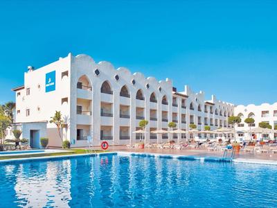 Großes Hotelgebäude mit weißen Wänden, blauer Fensterverkleidung und großem Schwimmbecken vor blauem Himmel.