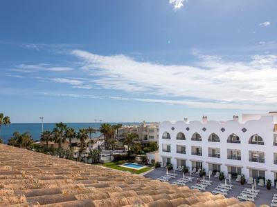 Blick auf Hotel mit weißen Gebäuden, Pool, Palmen und weiter Sicht auf das blaue Meer unter blauem Himmel.