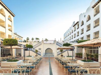 Cortile di un hotel con file di tavoli, sedie e grandi ombrelloni sotto un cielo blu.