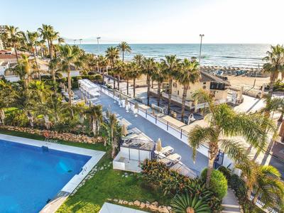 Blick auf Pool, Palmen und Fußweg neben Strand mit Meer im Hintergrund bei klarem Himmel.