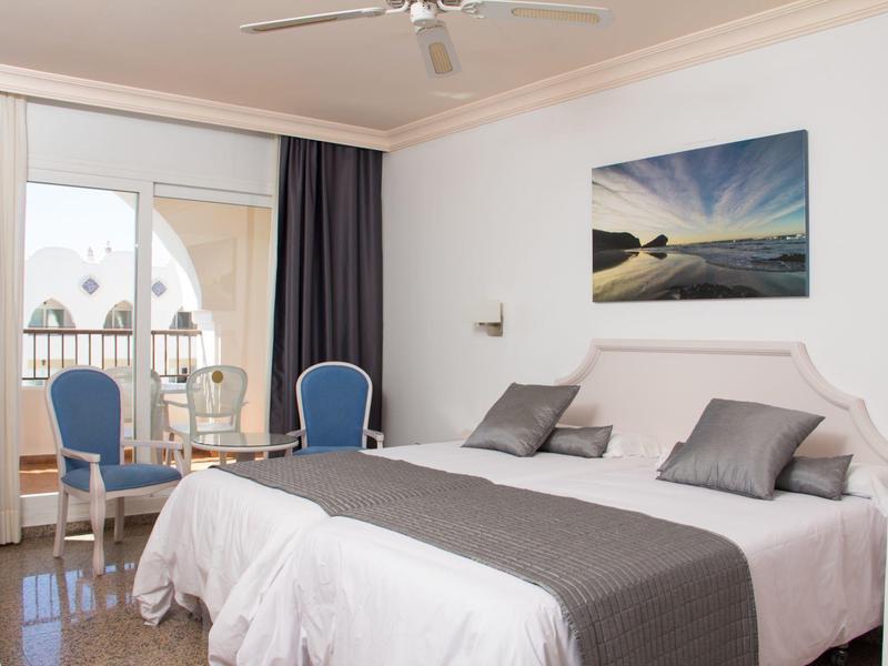 Helles Schlafzimmer mit Doppelbett, blauem Stuhl, Balkon und Meeresbild an der weißen Wand.