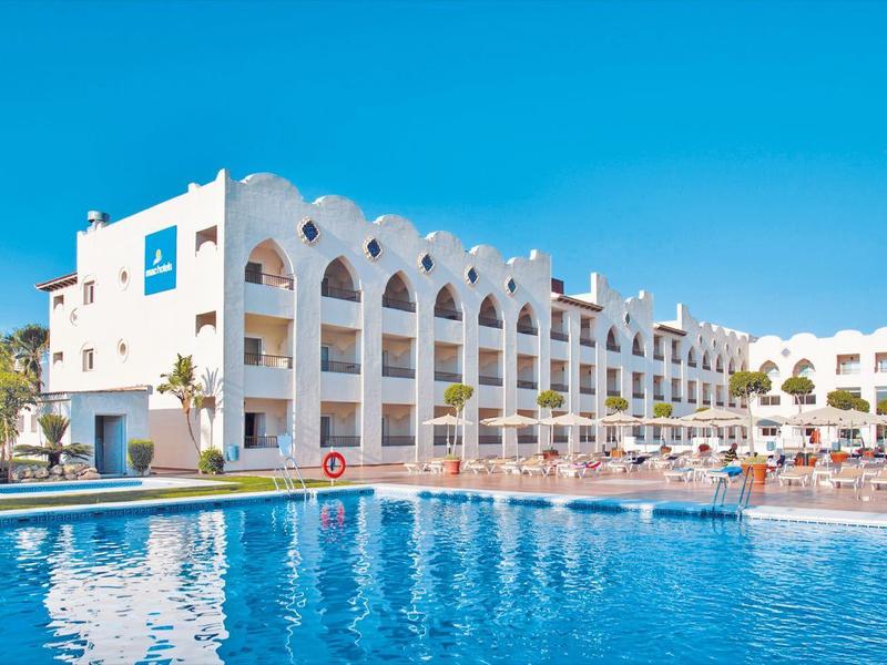 Großes Hotelgebäude mit weißen Wänden, blauer Fensterverkleidung und großem Schwimmbecken vor blauem Himmel.