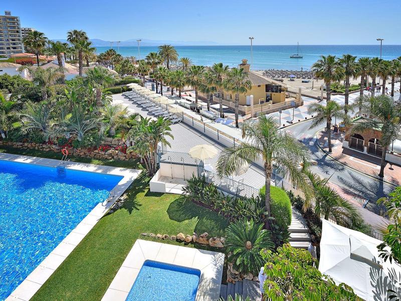 Blick auf Meer, Strand, Palmen und blauen Pool mit Liegestühlen in einer sonnigen Ferienanlage.