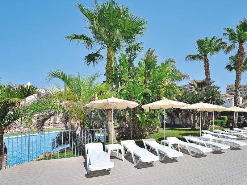 Poolbereich mit Liegestühlen, Sonnenschirmen und Palmen unter blauem Himmel im Freien.