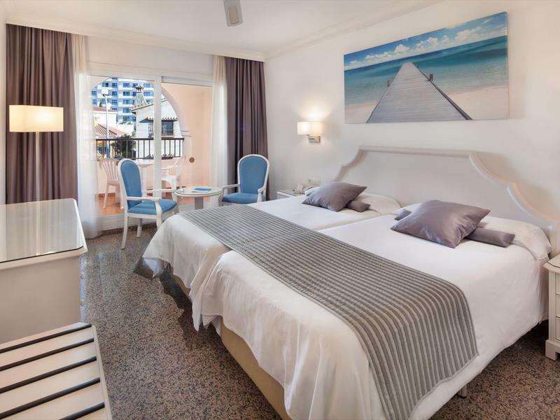 Helles Hotelzimmer mit zwei Betten, Sitzecke, Balkon und Landschaftsbild an der Wand.