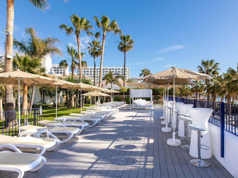 Strandpromenade mit weißen Liegen, Stroh-Sonnenschirmen und hohen Palmen unter blauem Himmel.