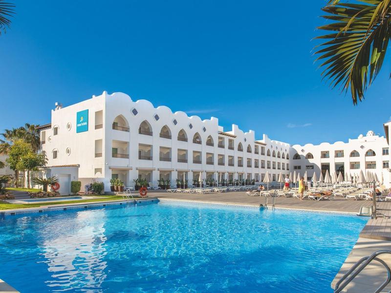 Großes Hotel mit weißer Fassade, mehreren Stockwerken und großem blauen Außenpool unter blauem Himmel.