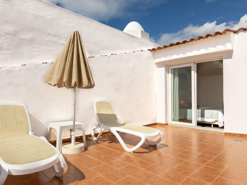 Terrasse mit zwei Liegestühlen, geschlossenem Sonnenschirm und weißen Wänden, blauem Himmel.