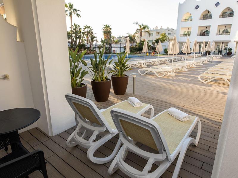 Blick von Balkon mit zwei Liegestühlen und Pflanzen auf Poolbereich vor einem weißen Gebäude.