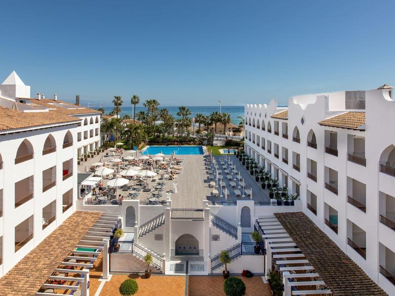 Großes Hotel mit weißer Fassade, Poolbereich und Blick auf das blaue Meer unter klarem Himmel.