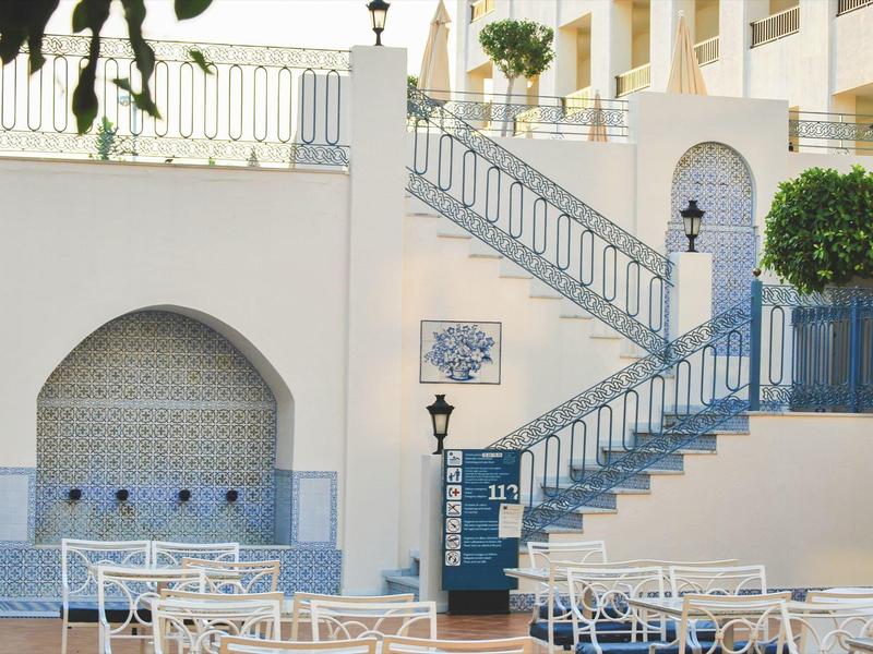 Terrasse mit weißen Tischen und Stühlen, Treppe mit blauem Geländer und wandmontiertem Brunnen.