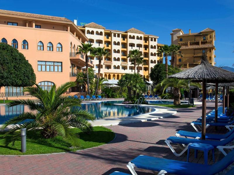 Hotelanlage mit Pool, Sonnenschirmen, Liegestühlen und Palmen unter klarem blauem Himmel.