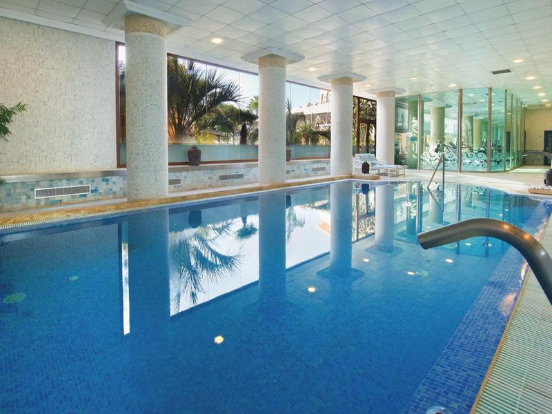 Großes Hallenbad mit blauen Fliesen, Säulen, großen Fenstern und moderner Beleuchtung.