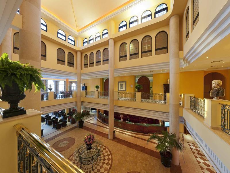 Helles, dreistöckiges Atrium mit Geländern, großen Fenstern, Pflanzen und orange-gelben Wänden.