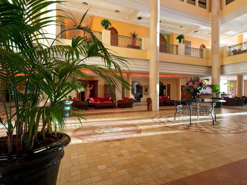 Helle Hotellobby mit großen Pflanzen, Säulen, Sitzgruppen und warmen Holz- und Beigetönen.