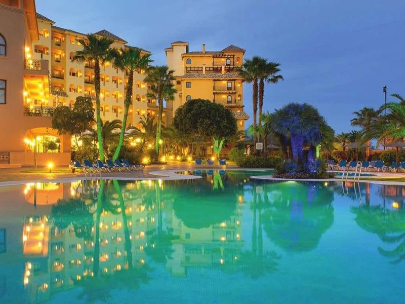 Großer Pool bei Dämmerung, umgeben von Palmen und beleuchteten Gebäuden in warmen Farben.