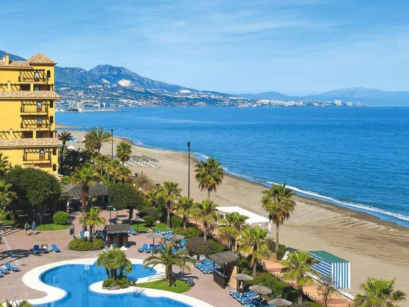 Strand mit blauem Meer, gelben Gebäuden und einem Poolbereich, dahinter Berge unter klarem Himmel.
