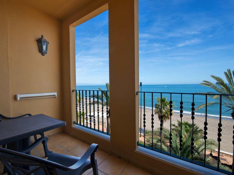 Balkon mit Stühlen und Tisch, Blick auf Meer, Palmen und blauen Himmel bei Tageslicht.
