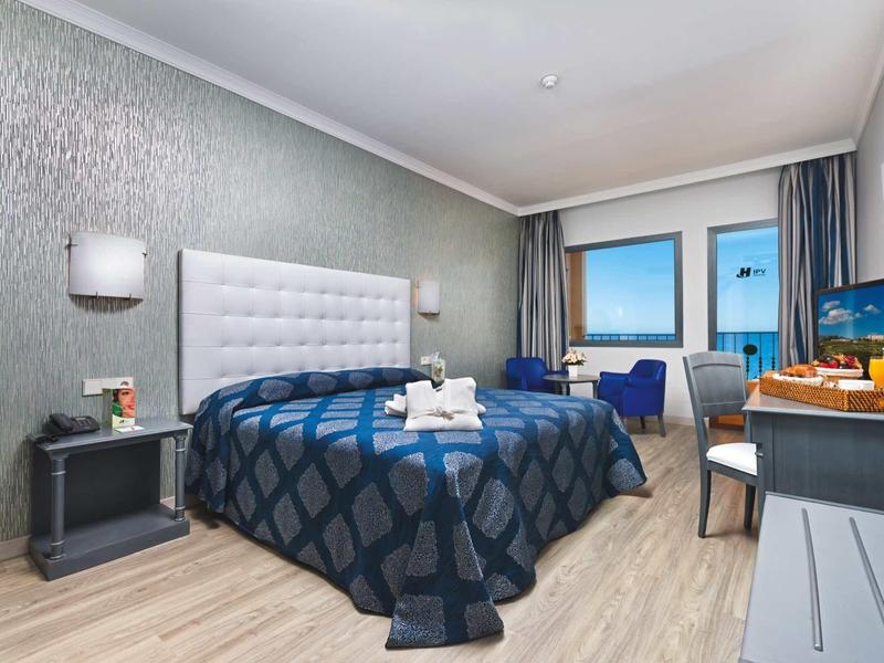 Modernes Hotelzimmer mit großem Bett, blauem Überwurf, Fenster mit Meerblick und Schreibtisch.